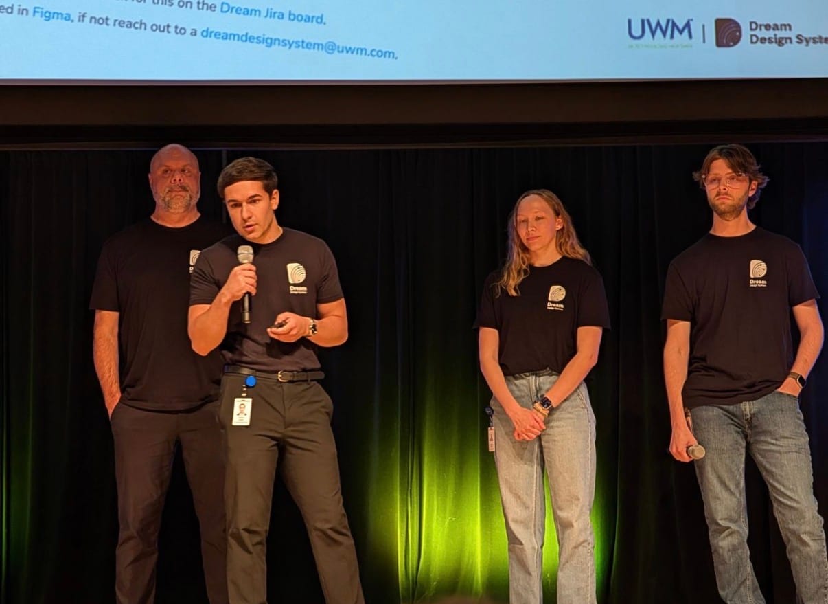 Aleks Duni speaking into a microphone on stage with three Dream team members at UWM's All-IT meeting